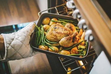 an overhead photo of a person reheating a chicken in oven in a pan surrounded by potatoes and asparagus