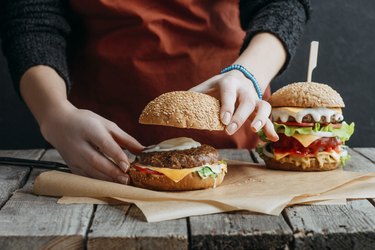 cropped view of girl in apron making homemade cheeseburgers on baking paper on wooden tabletop