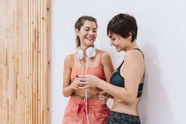 two friends women at the gym talking happily, listening to music and using smart phone. Technology, sports and friendship concept