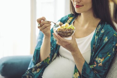 A smiling pregnant woman eating a bowl of granola topped with fruit