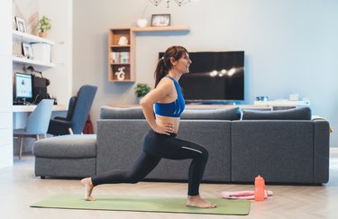 an adult wearing black leggings and a blue sports bra practices a lunge exercise at home on a green yoga mat