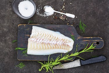 Raw cod before cooking on a black chopping Board with herbs and sea salt on a dark background. Top view