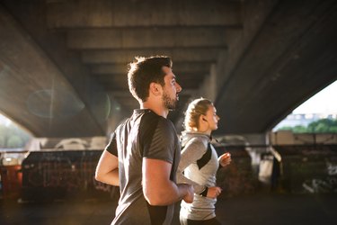 Young sporty man and woman with earphones running under the bridge outside in a city.