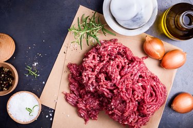 Minced ground beef on craft paper with onions, herbs and spices on side