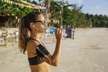 Woman drinking water on the beach and listening to her summer workout playlist