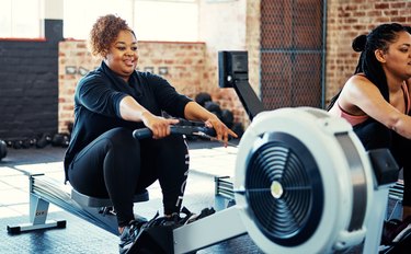 Woman in black leggings and a black workout jacket on a rowing machine, doing a 20-minute workout, in a brick-walled gym, next to another woman also on a rowing machine