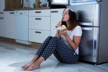 person wearing blue polka dot capri pants and a white t-shirt sitting on floor next to refrigerator eating a piece of cake and savoring a bite with fork in mouth
