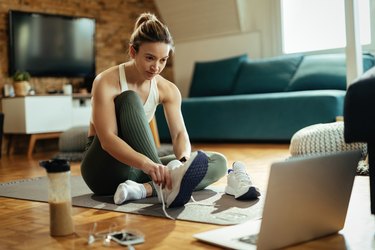 Athletic woman putting on sneakers while using laptop on the floor.