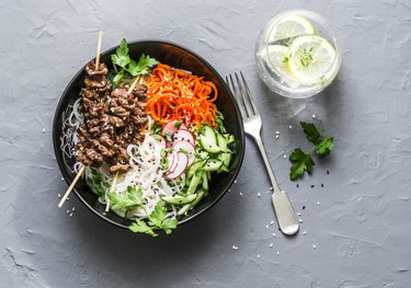 Rainbow power buddha bowl. Asian style beef skewers, rice vermicelli, pickled vegetables salad carrots, cucumbers, radishes, greens and lemon thyme lemonade on grey background, top view