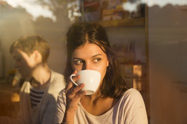 Woman drinking coffee in urban cafe while looking outside, sunny reflections in window.