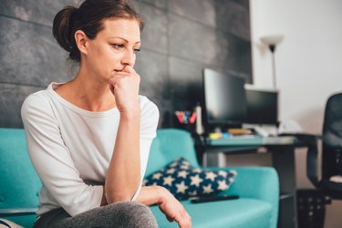 a person looks anxious sitting on a teal couch at home with a desk and a computer in the background