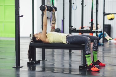 Athlete wearing yellow tank top and black leggings performing a bench press with dumbbells in gym.