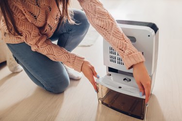 a close up photo of a person wearing a peach long-sleeve sweater and jeans changing the water container of a dehumidifier at home