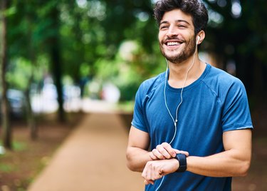 A smiling man wearing headphones and checking his smartwatch after a jog in the park