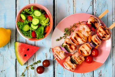 Grilled chicken and vegetable skewers with salad and watermelon, above scene over blue wood