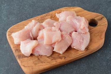 Raw chicken fillet cut in big cubes on a olive wood cutting board on a grey abstract background. Step by step cooking