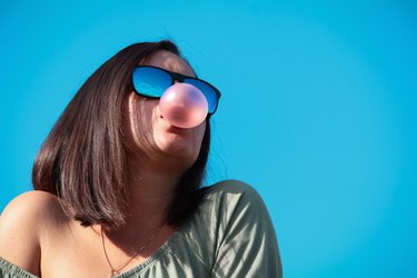 Low Angle View Of Woman Wearing Sunglasses Chewing Gum Blowing a Bubble Against Blue Background