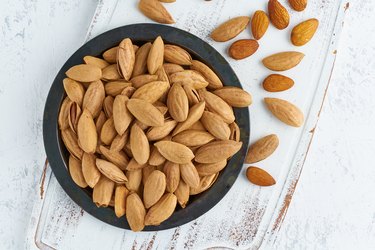 Top view plate with almonds in endocarp, bowl with drupe in shell on a white wooden cutting board. White background, overhead, copy space.