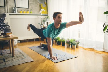 man balancing on one knee and one hand practicing yoga on mat at home
