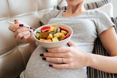 Pregnant young woman sitting and eating fruit salad at home