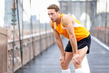 a runner wearing a yellow singlet, black shorts and white knee-high socks rests on the Brooklyn Bridge with their hands on their knees