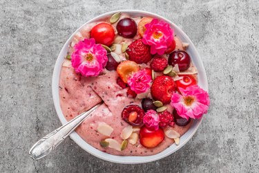 bowl of pink berry smoothie or nice cream made of frozen fruits and nuts with fresh berries, nuts and rose flowers