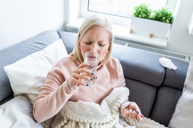 Concept of suffering from terrible pain. Beautiful, sad, upset, unhappy, troubled, weak woman with blonde hair, wearing casual clothes is sitting on a sofa and taking a pill