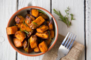 A bowl of hearty, beta-carotene-rich sweet potatoes with fresh rosemary sprigs on a white wooden table.