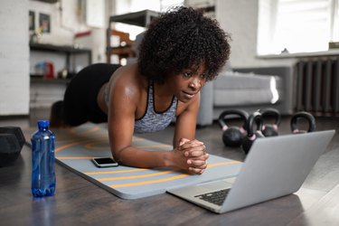 woman doing plank exercise in sports bra and leggings in front of laptop at home