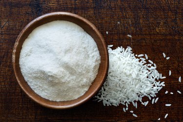 White rice flour in dark wooden bowl isolated on dark brown wood from above. Spilled rice.