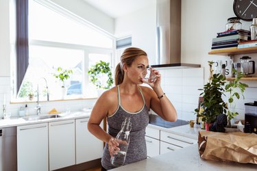 穿着运动服的女人站在家里的厨房里喝水