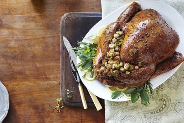 an overhead photo of a whole cooked turkey with glaze, herbs and stuffing on fresh herbs and lemons on a white serving platter with carving utensils on a wooden table