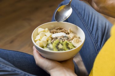 an overhead photo of a person holding a white bowl of yogurt, buckwheat, seeds and fresh fruit and a spoon