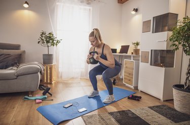 Young woman squatting with kettlebell on yoga mat in living room