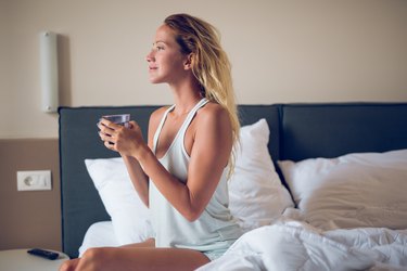 A woman drinking a glass of water while sitting on her bed