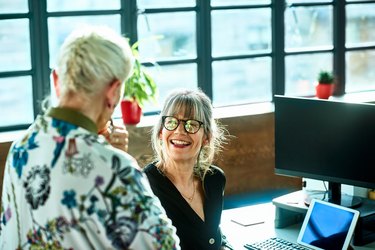 Cheerful mature woman at desk smiling towards senior female colleague