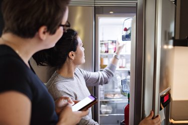 Woman with mobile phone looking at girlfriend opening refrigerator in kitchen