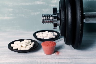 Creatine Powder, Aginine capsules, BCAA (amino acids) and a dumbbell in background. Sport nutrition. Wooden background. Close up.