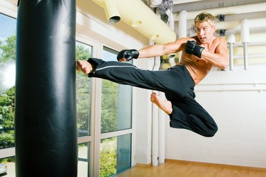 A topless man jumping up and kicking a heavy kicking bag