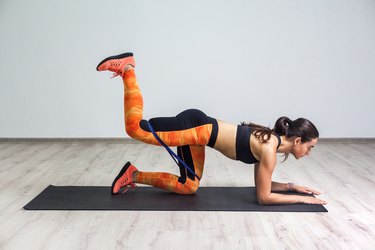 Side view portrait of strong and fit athletic young woman in sportswear with bands training legs and glutes muscular. Fitness exercising with expander.