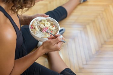 an overhead photo of a person wearing a black tank top and leggings eating a bowl of oatmeal after a workout sitting on a wooden floor