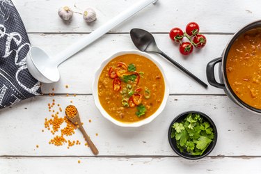 Bowl and pot of garnished red lentil soup and ingredients on white wood