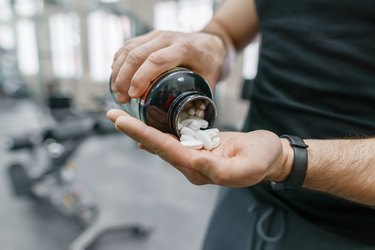Closeup of sporty muscular arms in a black shirt and pants pouring white supplements into the palm of hand.