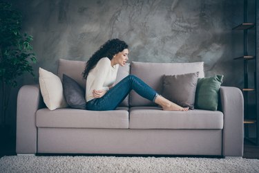 Profile side view of her she attractive depressed unhealthy wavy-haired girl sitting on divan suffering from medical belly problem in modern loft industrial style interior living-room indoors