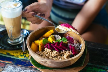 Woman's hands with smoothie bowl made with mango, banana, granola, grated coconut, dragon fruit and mint with latte coffee for plant based breakfast