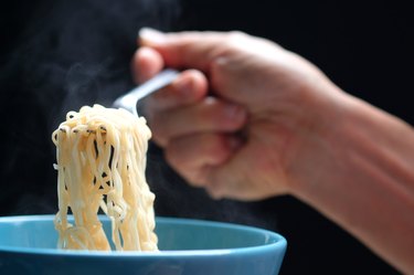 Hand holding fork full of hot noodles from soup in a blue bowl.