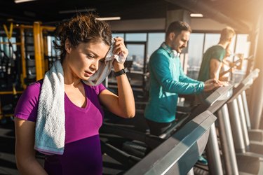 Young tired woman wiping sweat with towel while running on a treadmill in a gym