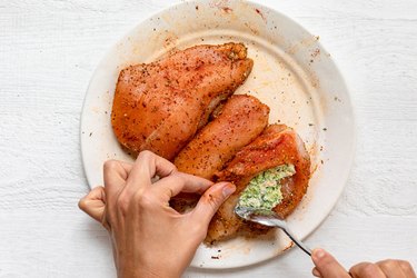 A hand uses a spoon to stuff a broccoli-cheese mixture into a seasoned chicken breast