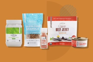 Five of the staples included in Thrive Market's staples kit on an orange background: Coconut oil, grain-free granola, pink salt, beef jerky and canned salmon