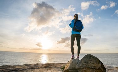 woman wearing backpack standing on rock on beach at sunset